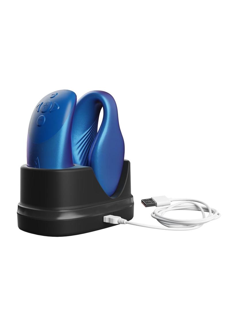A blue couples vibrator with an interactive squeeze remote control. This image includes the discreet charging dock. The charging station is black. Easily connect the charging station USB-C to a USB brick charger to power on. The vibrator has a clamshell c-shaped curved design, with parts that vibrate externally on the clit and internally on the g-spot. The remote is sleek, small enough to fit in the palm of your hand. The remote has buttons to increase and decrease intensity as well as change the pattern. 