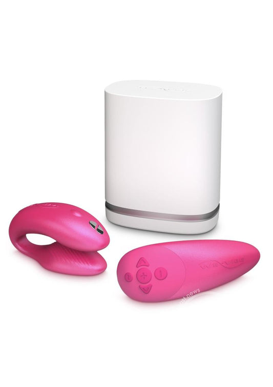 A pink couples vibrator with an interactive squeeze remote control. This image includes the discreet charging dock. The charging station is white, with a lid that says "We Vibe." The vibrator has a clamshell c-shaped curved design, with parts that vibrate externally on the clit and internally on the g-spot. The remote is sleek, small enough to fit in the palm of your hand. The remote has buttons to increase and decrease intensity as well as change the pattern. This toy is hot pink in color.